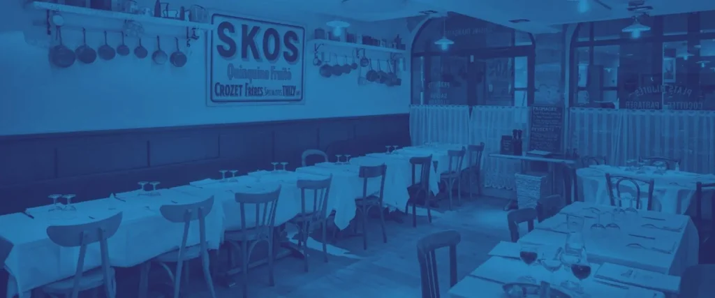 Vue panoramique de la salle du Bistrot Marie avec un filtre bleu esthétique, montrant les tables dressées, les casseroles en cuivre et l'affiche vintage SKOS