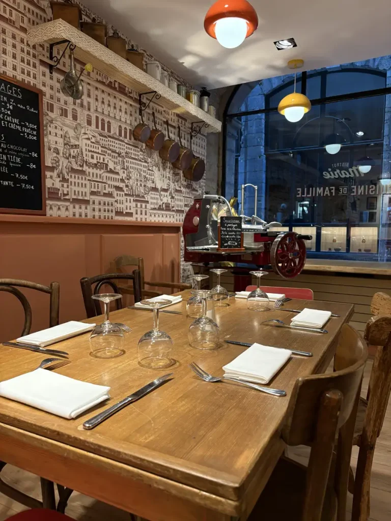 OUVERTURE FÉRIÉS 4 Table en bois dressée avec verres à vin et serviettes blanches, trancheuse à saucisson rouge et ardoise au Bistrot Marie Vieux-Lyon